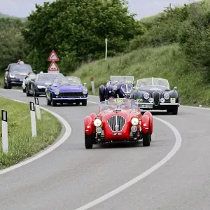 Several classic cars in a convoy drive on a winding country road through a green landscape Several classic cars in a convoy drive on a winding country road through a green landscape