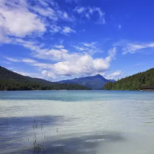 A clear lake with surrounding mountains and wooded slopes under a blue sky with white clouds