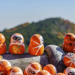 Close-up of Daruma figures with an autumnal mountain in the background and clear sky Close-up of Daruma figures with an autumnal mountain in the background and clear sky