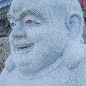 Closeup of head and face of stone carved sitting Buddha with blurred temple building in background in Pyeongtaek