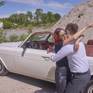 A couple embracing joyfully by a white convertible car on a sunny day next to building site of work