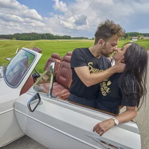 Couple kissing by a convertible car amidst a rustic countryside backdrop