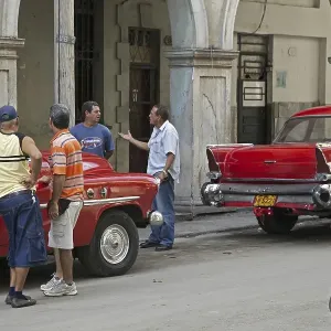Cuban men discussing on street and red old 1950s vintage American cars