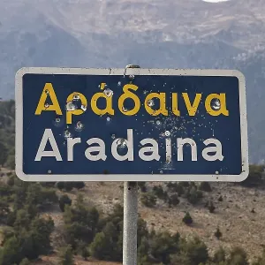 Damaged road sign with Greek and Latin lettering in front of a mountain landscape Damaged road sign with Greek and Latin lettering in front of a mountain landscape