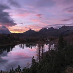 Ein ruhiger Bergsee, umgeben von Waldern und Bergen wahrend des Sonnenaufgangs mit rosa und lila Wolken