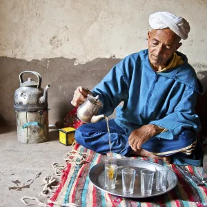 Elderly Berber man wearing a turban sitting on the floor on a rug pouring traditional mint tea from a silver jug Elderly Berber man wearing a turban sitting on the floor on a rug pouring traditional mint tea from a silver jug