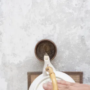 Hand gracefully pours tea from a white ceramic teapot with bamboo handle into a small brown bowl on a textured gray Hand gracefully pours tea from a white ceramic teapot with bamboo handle into a small brown bowl on a textured gray