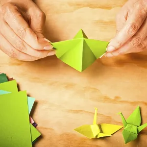 Hands working on green origami crane with colored paper nearby
