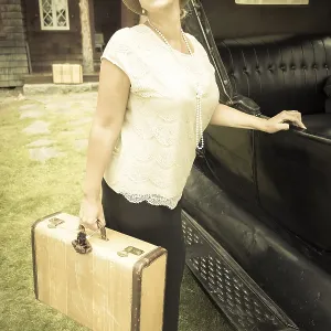 Happy 1920s dressed girl holding suitcase next to vintage car and cabin Happy 1920s dressed girl holding suitcase next to vintage car and cabin