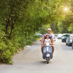 Happy young father and little daughter riding a vintage scooter in the street wearing hats
