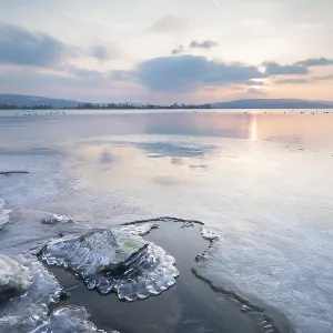 Icy coast with rocks, sunset over a quiet lake with mountain landscape Icy coast with rocks, sunset over a quiet lake with mountain landscape