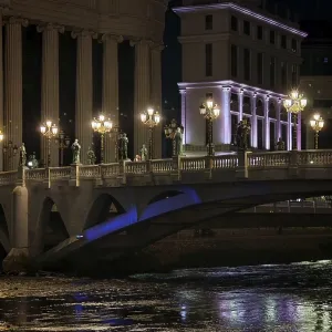 Illuminated bridge over a river at night with urban architecture in the background