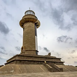 An imposing lighthouse rises against a cloudy sky surrounded by historic architecture