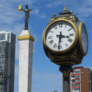 Large clock and statue in front of modern buildings under bright blue sky