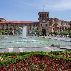 Large square with fountain, surrounded by flower beds and a historic building with clock