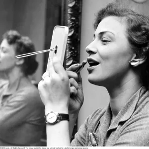 Looking good in the 1950s. She is looking at herself in the pocket mirror while applying lipstick