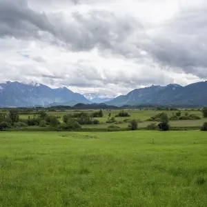Pradera verde y espesa que se extiende hacia montañas boscosas bajo un cielo nublado dramático.