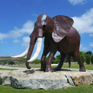 Metal sculpture of an elephant on a rock foundation in a green park under a blue sky with white clouds Metal sculpture of an elephant on a rock foundation in a green park under a blue sky with white clouds
