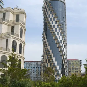 A modernly designed high-rise building with spiral balconies rises above the surrounding green areas