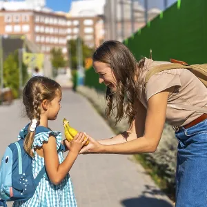 Mother smiles as she hands her schoolgirl a banana on a sunny walk