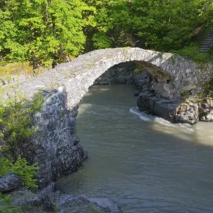 Old stone bridge spanning river in a green forest area in sunshine