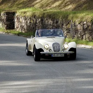 An old-timer drives quickly along a winding road with a stone wall