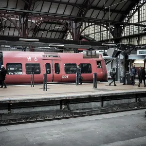 Passengers near a red train at a train station with urban architecture in Copenhagen Passengers near a red train at a train station with urban architecture in Copenhagen