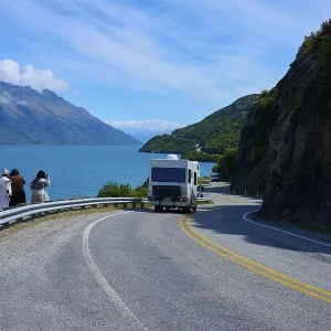 People enjoying travelling and a motorhome on a winding road next to a lake surrounded by mountains