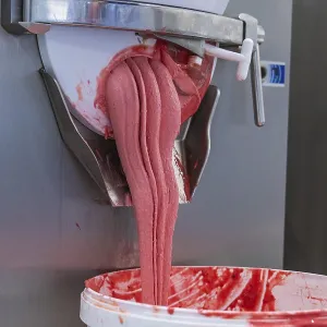 Pink-coloured ice cream mass flows viscously from a machine into a bucket