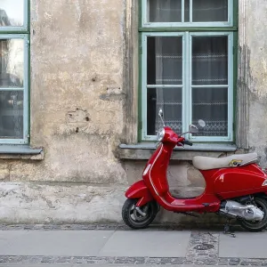 A red vintage scooter on a pedestrian sidewalk in front of an old stone house