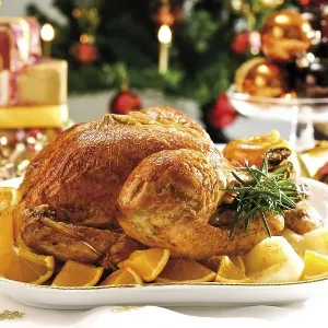Roast turkey surrounded by oranges in front of wrapped presents and a decorated Christmas tree