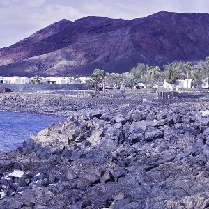 Rugged coastal area with rocks and views of reddish brown mountains under cloudy sky