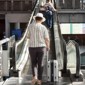 Senior woman in white hat pulling suitcase up an escalator inside modern transport terminal