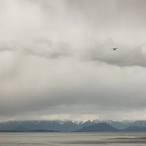 A small airplane flies over a wide area with distant mountains as seen from deck of passenger ferry crossing the Salish A small airplane flies over a wide area with distant mountains as seen from deck of passenger ferry crossing the Salish