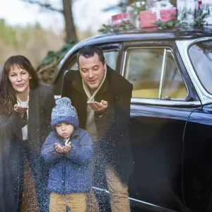 Smiling adult man and woman with boy in warm clothes blowing snow standing at vintage car with Christmas presents Smiling adult man and woman with boy in warm clothes blowing snow standing at vintage car with Christmas presents