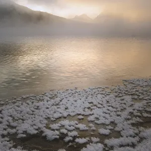 Snow-covered lake shore with distant misty mountains under a golden sunrise