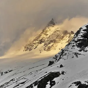 A snowy mountain peak rises majestically through dramatic clouds at twilight A snowy mountain peak rises majestically through dramatic clouds at twilight