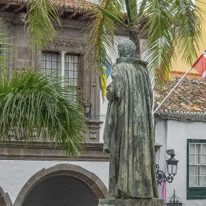 Statues and palm trees in front of colonial building with flags in a square