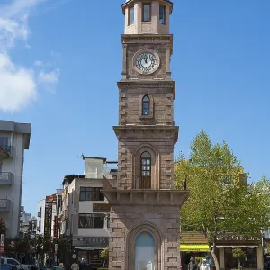 A tall clock tower in an urban square with trees and shops
