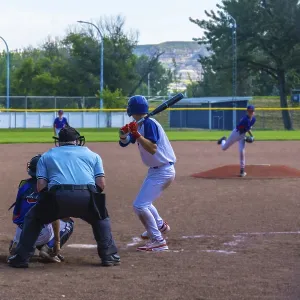 Teenage baseball batter waiting for the pitch while the pitcher is throwing the ball during a sunny summer game