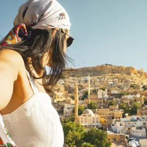 Traveler woman in white dress arms taking selfie self-portrait ooking over panoramic view of historic city of Mardin