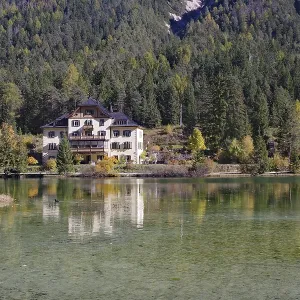 The villa is brilliantly reflected in a quiet alpine lake in front of dense forest and gentle slopes