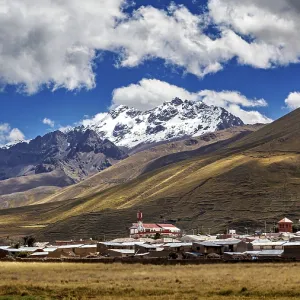 A village at the foot of snow-capped mountains under a blue sky with clouds