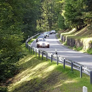 Several vintage cars drive on a winding country road through a sunny forest