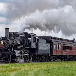 A vintage steam train chugs along railway tracks surrounded by lush green fields under a clear blue sky