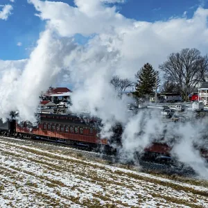 A vintage steam train moves through a landscape of snow-dusted fields under a clear blue sky