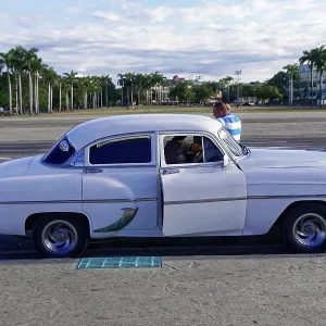 White vintage car in front of palm tree-lined square with people in relaxed surroundings