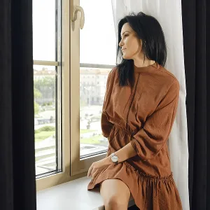 A woman in a brown dress relaxes by a large window