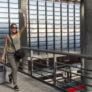 Woman smiling and holding a ticket while walking with her luggage through a futuristic transportation hub