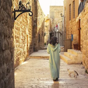 Woman in traditional abaya walking with cat through narrow stone alley in historic city of Mardin Turkey with ancient